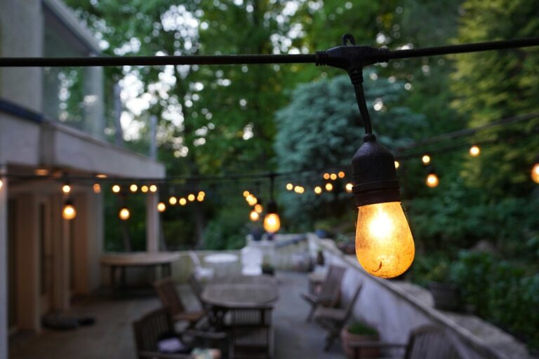 Warm outdoor lights illuminate a patio, garden path, mature trees, and pool deck at dusk.
